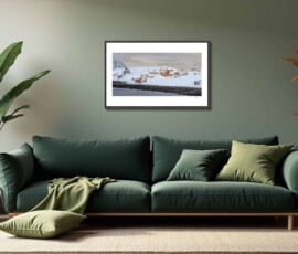 Fine-art photography print titled Arctic winter Sarnesfjorden I by Eva Eidsheim Photography. Showing a color cultural landscape of a tiny coastal settlement in Nordkapp on an icy winter day. Waves crash against the shore of a snow-covered landscape under a dark sky, capturing a raw Arctic atmosphere, printed on premium Hahnemühle art paper.